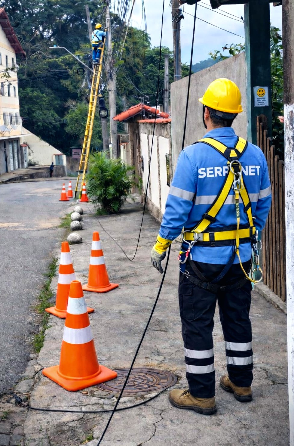 Técnico da SERRANET de uniforme e cinto de segurança instalando fibra óptica num poste, em uma rua da serra fluminense